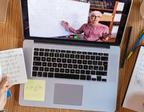 A high-angle view of a student's laptop on a desk covered in notes. On the screen, an instructor tecahes mathematics. 