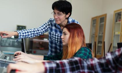 student on computer in foreground with teacher helping another student in background
