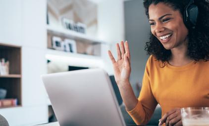 A young African-American woman smiles and waves at her laptop screen as she attends a webinar.