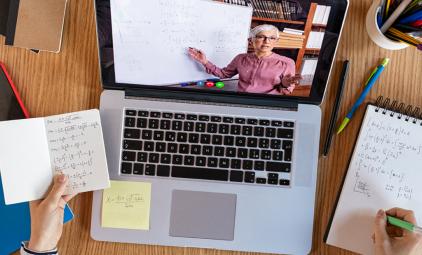 A high-angle view of a student's laptop on a desk covered in notes. On the screen, an instructor tecahes mathematics.