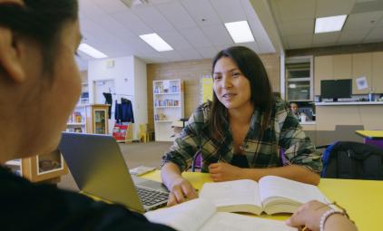 Student at a library table with open books and a laptop