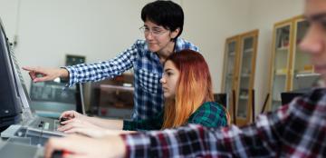 student on computer in foreground with teacher helping another student in background