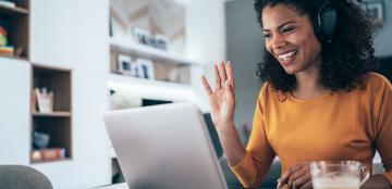 A young African-American woman smiles and waves at her laptop screen as she attends a webinar.