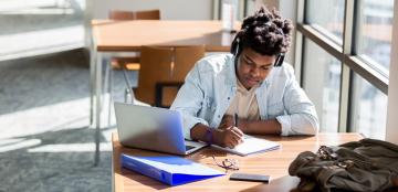 A male African-American student studies at a table in a brightly-lit campus room. 