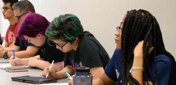 A group of college-aged students sits together at a table in a mathematics classroom. 