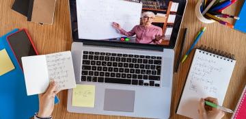 A high-angle view of a student's laptop on a desk covered in notes. On the screen, an instructor tecahes mathematics. 