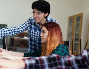 student on computer in foreground with teacher helping another student in background