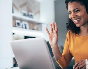 A young African-American woman smiles and waves at her laptop screen as she attends a webinar.