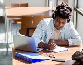 A male African-American student studies at a table in a brightly-lit campus room. 