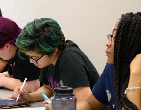 A group of college-aged students sits together at a table in a mathematics classroom. 