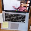 A high-angle view of a student's laptop on a desk covered in notes. On the screen, an instructor tecahes mathematics. 